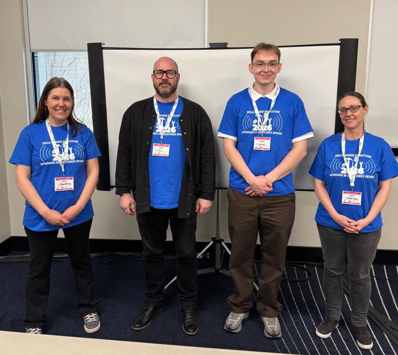 Four adults standing in front of a projector screen in a line &ndash; all wearing matching blue t-shirts and white and red lanyards. 