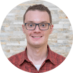 Chris Harlow, a man with light skin, glasses, short blonde hair and a red checked collared shirt, smiles against a brick background.