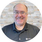 Andr&eacute; Piasta, a man with light skin, glasses, and a gray Esri Canada polo shirt, smiles against a brick background.