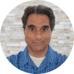 Nigel Pereira, a man with brown skin, wavy salt-and-pepper hair and a blue checked collared shirt smiles against a brick background.