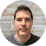 Ray Caron, a man with light skin, short dark hair, and a gray sweater stands against a brick background.