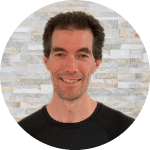Oli Helm, a man with light skin, short curly hair and a black crew-neck shirt, smiles against a brick background.
