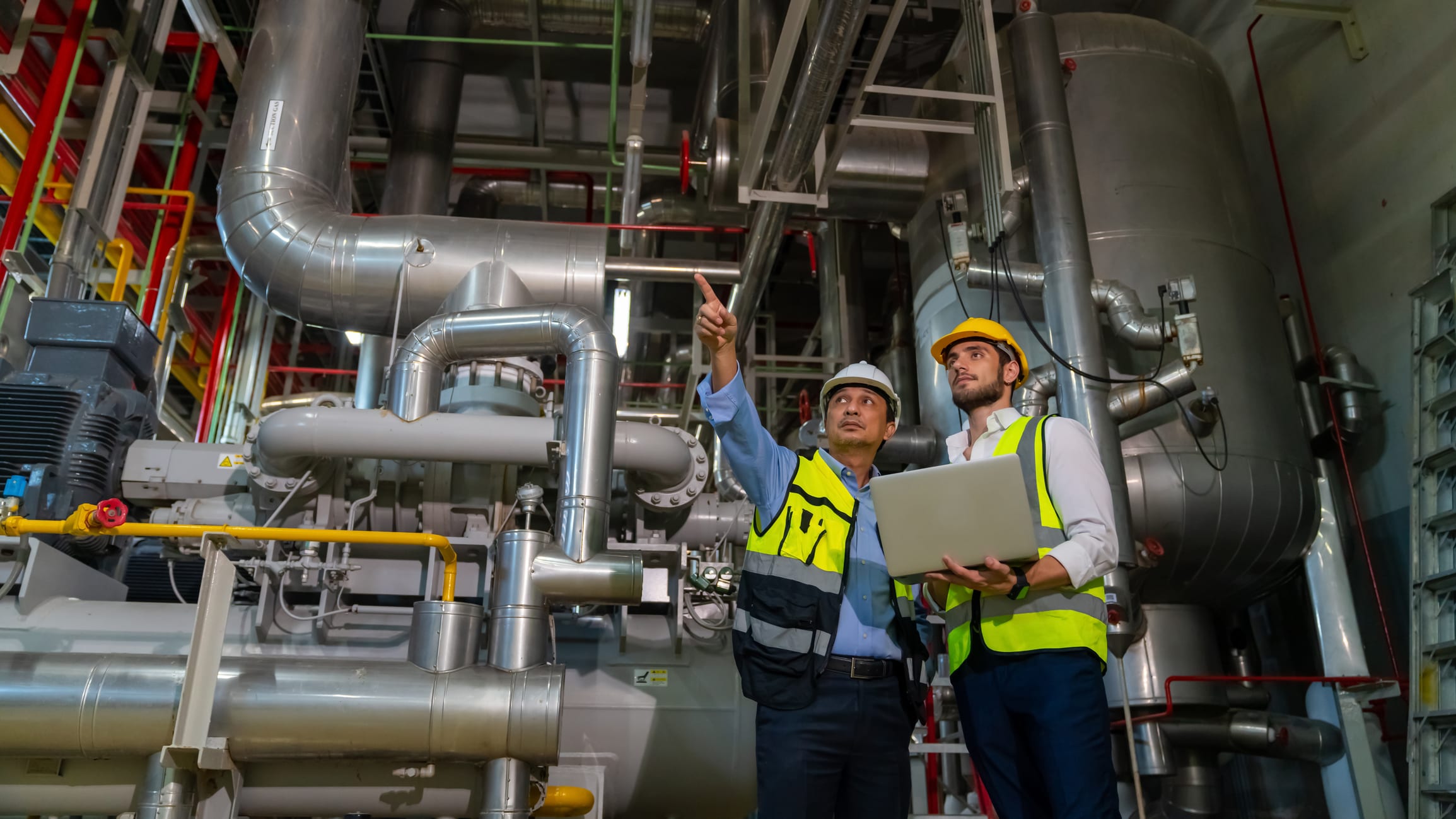Two workers in safety gear standing in an industrial plant, pointing at large pipes and machinery.