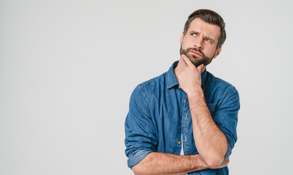 Against a white backdrop, a light-skinned man with a dark beard and dark hair and wearing a blue shirt holds his chin in confusion.
