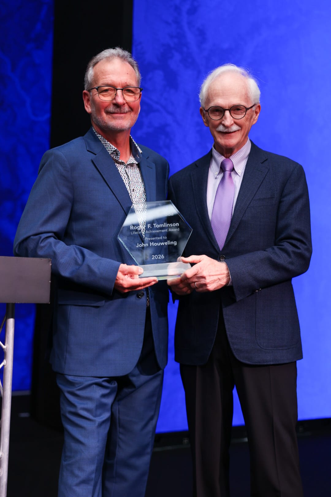 Two men in suits smiling on stage; one holds a glass plaque.