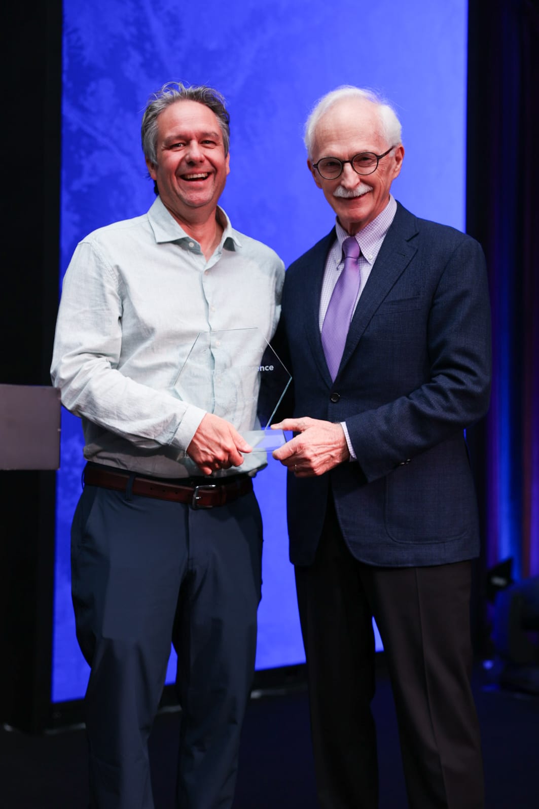 Two men smiling on stage; one holds a glass plaque.