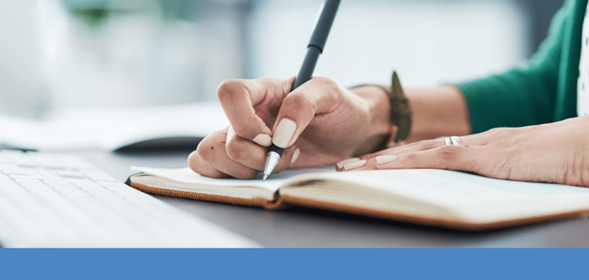 Woman's hands writing in a notebook laying in front of a laptop.