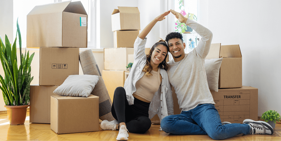 Boxes behind a couple sitting on the floor joining arms to make the shape of a house.