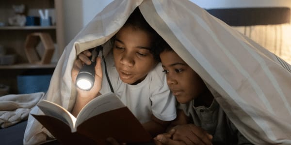Girl Reading mystery Book To Younger Brother with flashlight