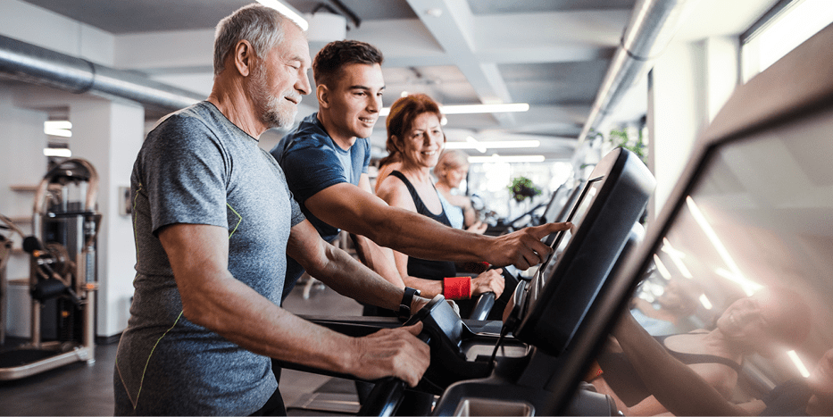 Younger man helping older man use treadmill while older woman watches.