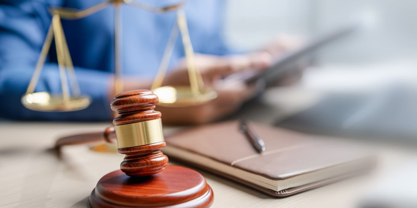 A gavel sitting in front of a notebook, pen, laptop, and a person holding a tablet.