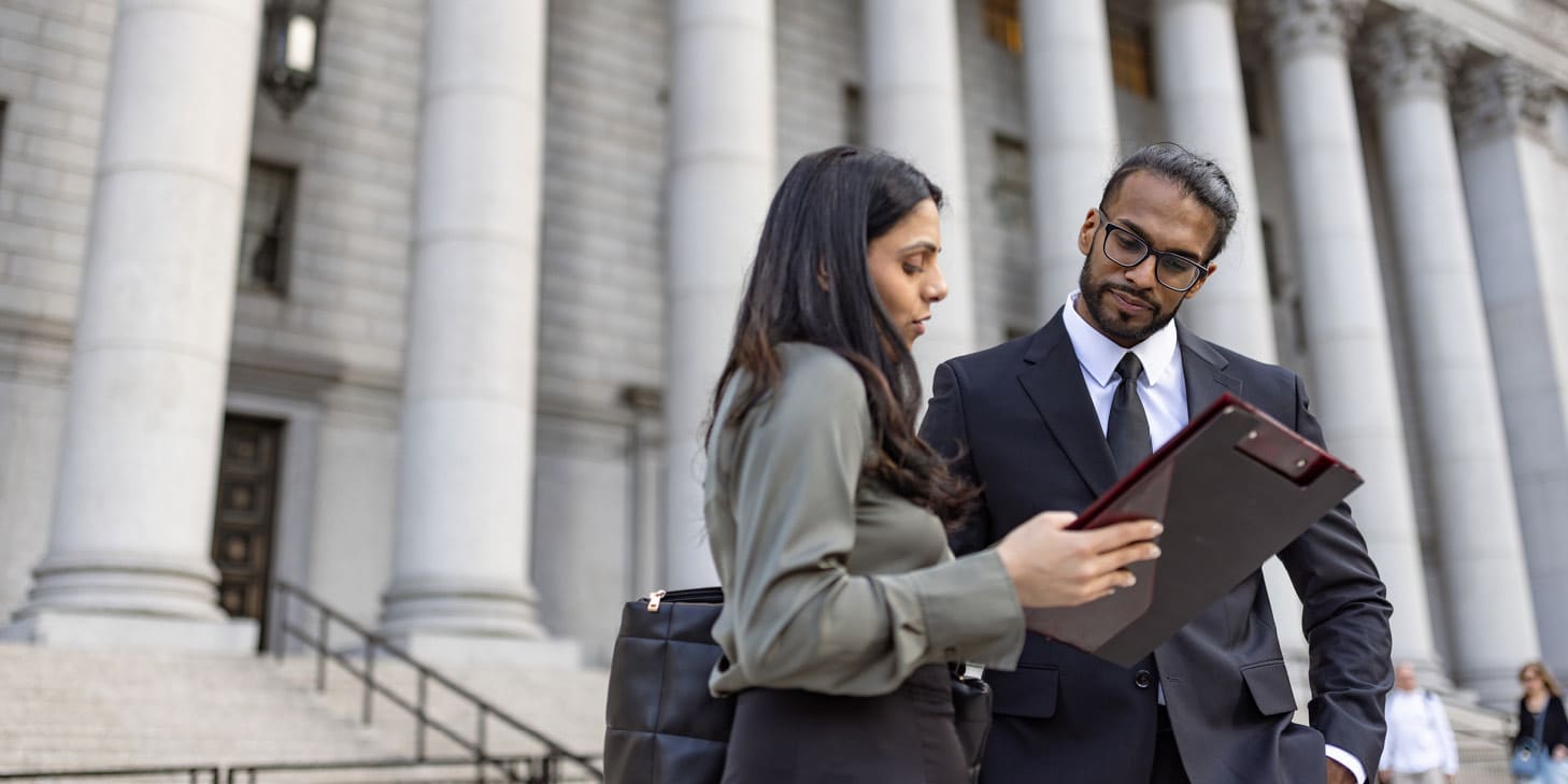 Two professionals, a man and a woman, reviewing documents on a tablet in front of a courthouse.