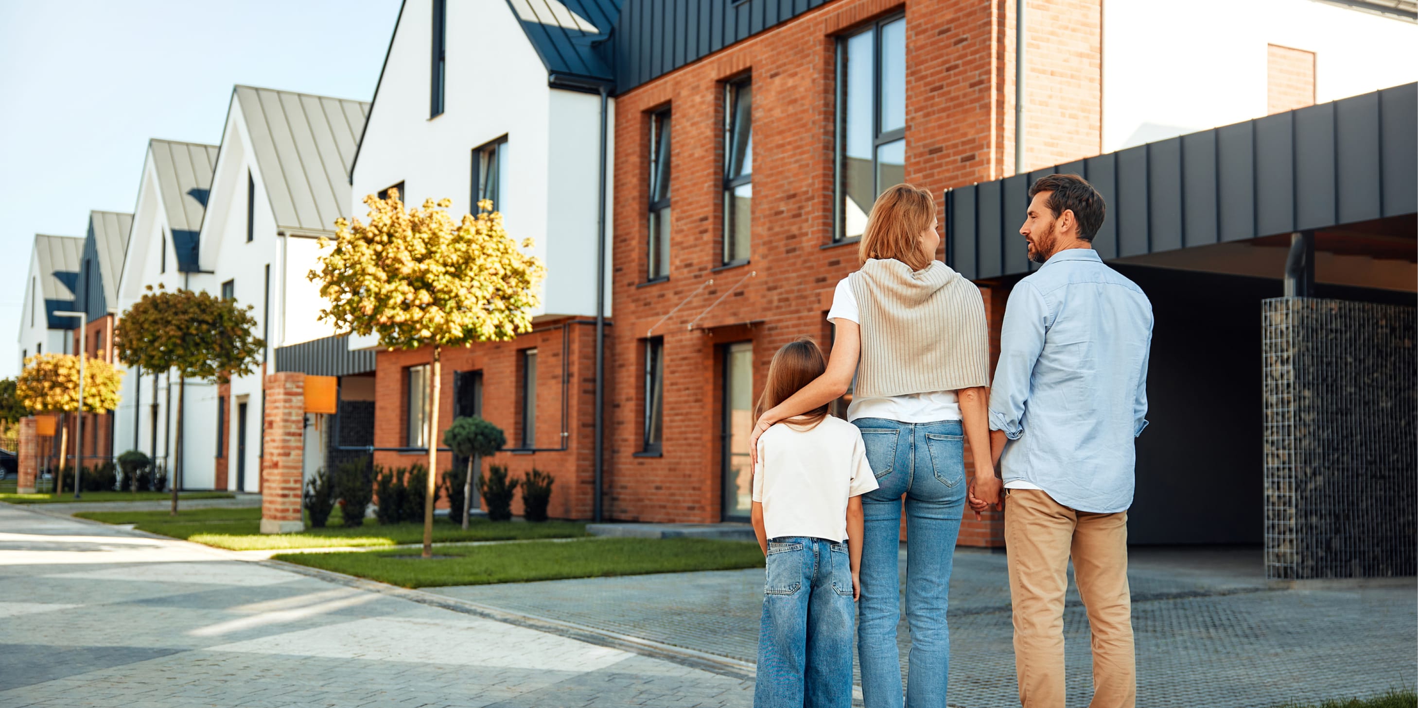 A family of three with a young daughter stands on a street and hugs each other in front of their home.