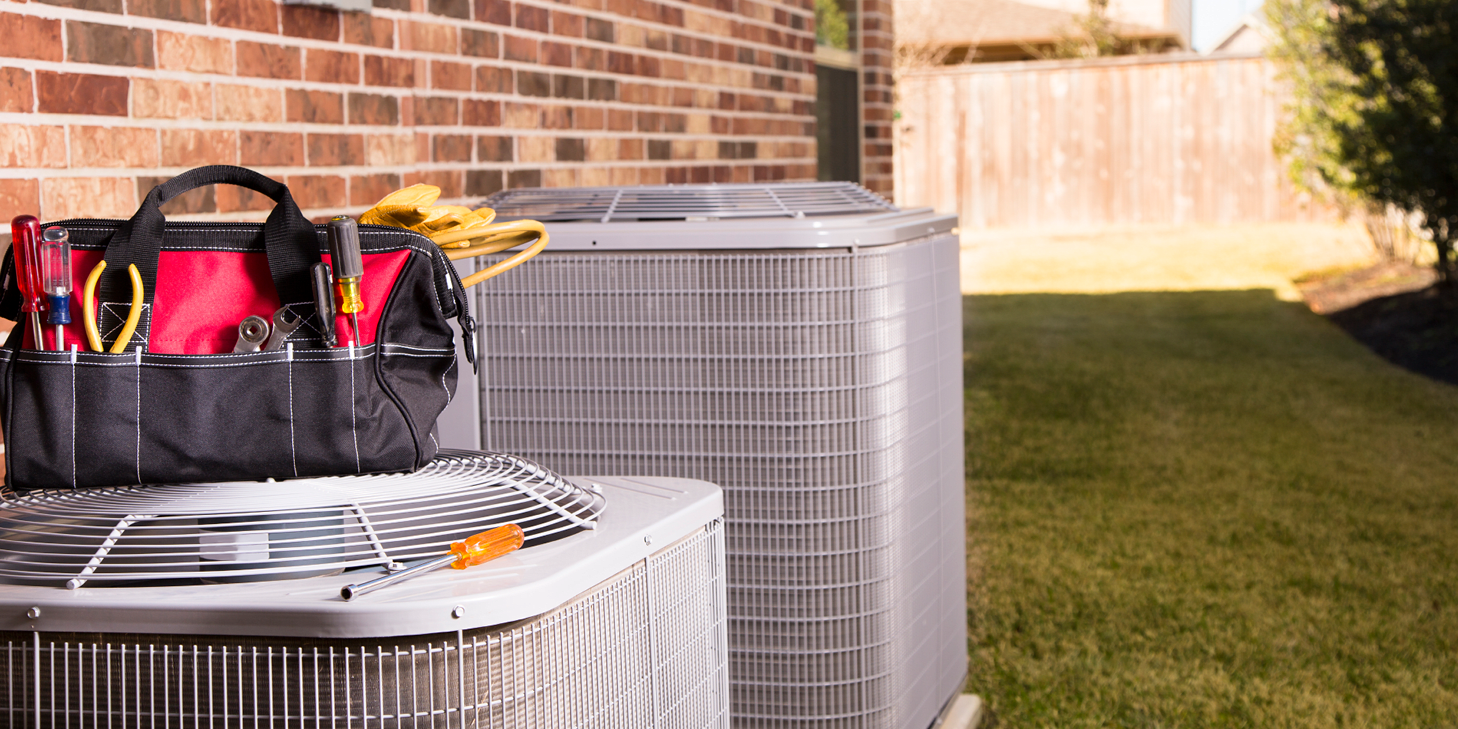 A bag with a repairman's work tools and gloves on top of two air conditioner units outside a brick home in a residential neighborhood.