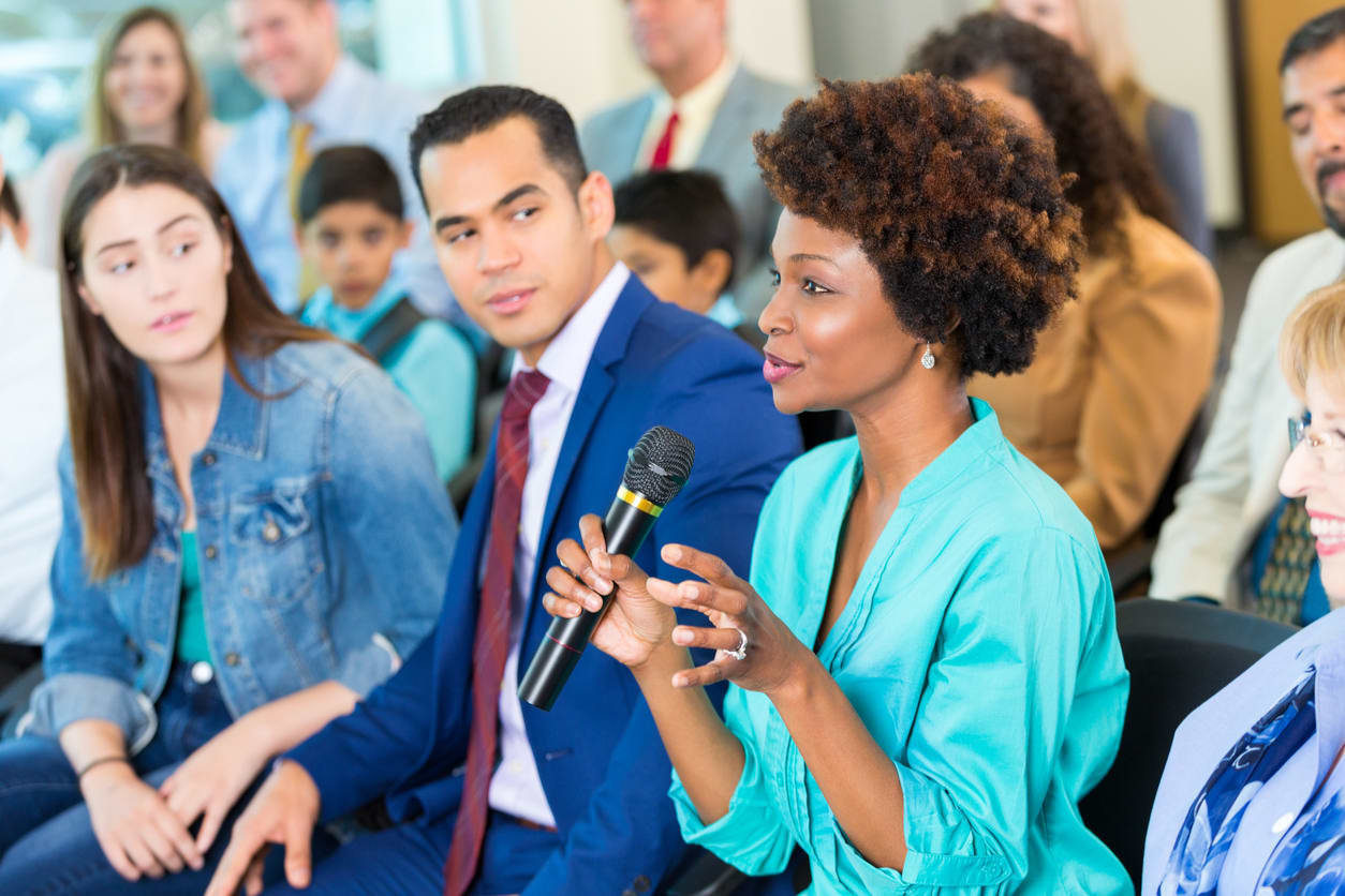 Woman speaking holds a microphone while seated in a community meeting.