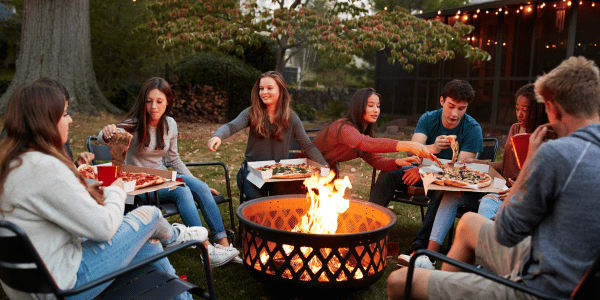 group of teenagers gathered around an outdoor fire pit eating pizza