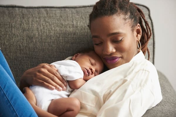 A Black mother resting on a couch cradles her sleeping infant