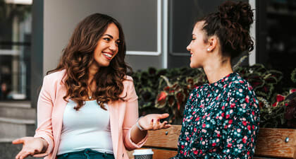 Two women smile as they talk over coffee outside.