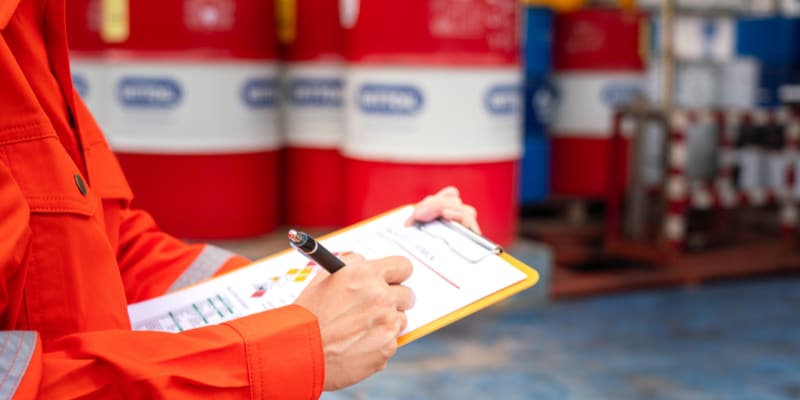 Image of an employee with a clip board standing in front of a barrel of hazardous materials.