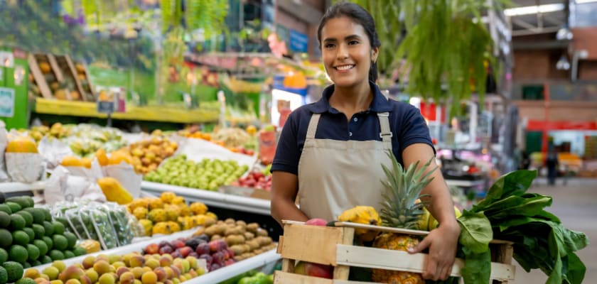 Grocery worker holding a basket of fruits and vegetables at the supermarket while smiling.