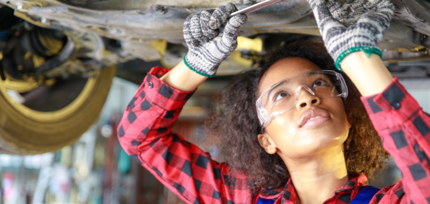 Mechanic wearing safety glasses working on a car on a lift above her.