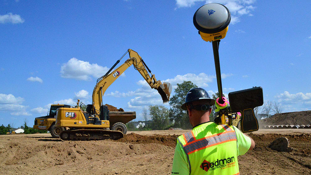 Worker carries survey equipment with machines in background