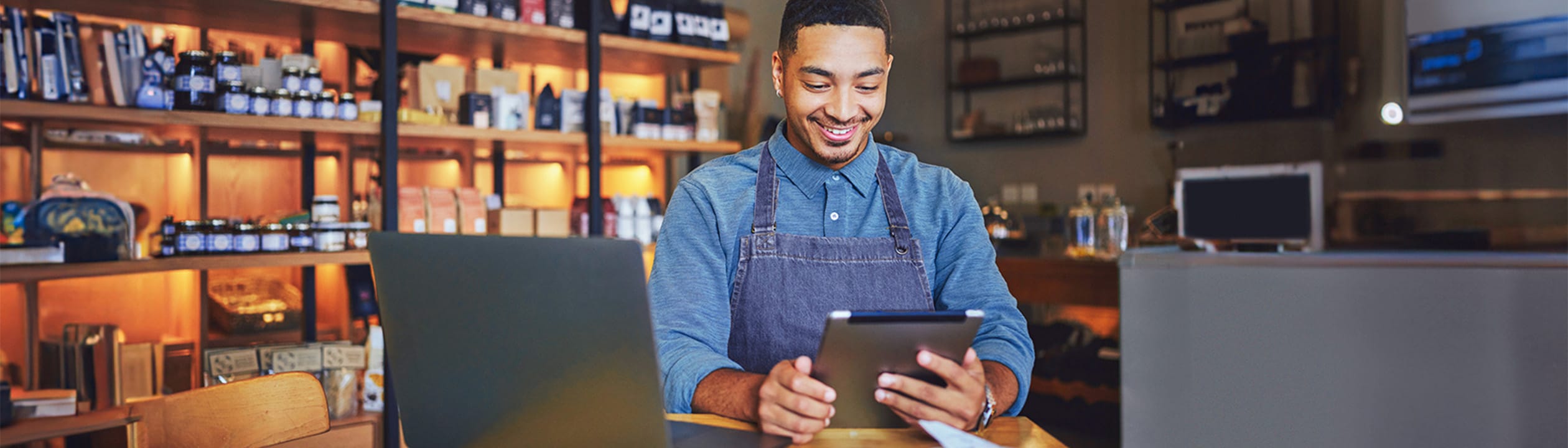 A smiling barista in an apron using a tablet at a coffee shop counter with a laptop nearby.