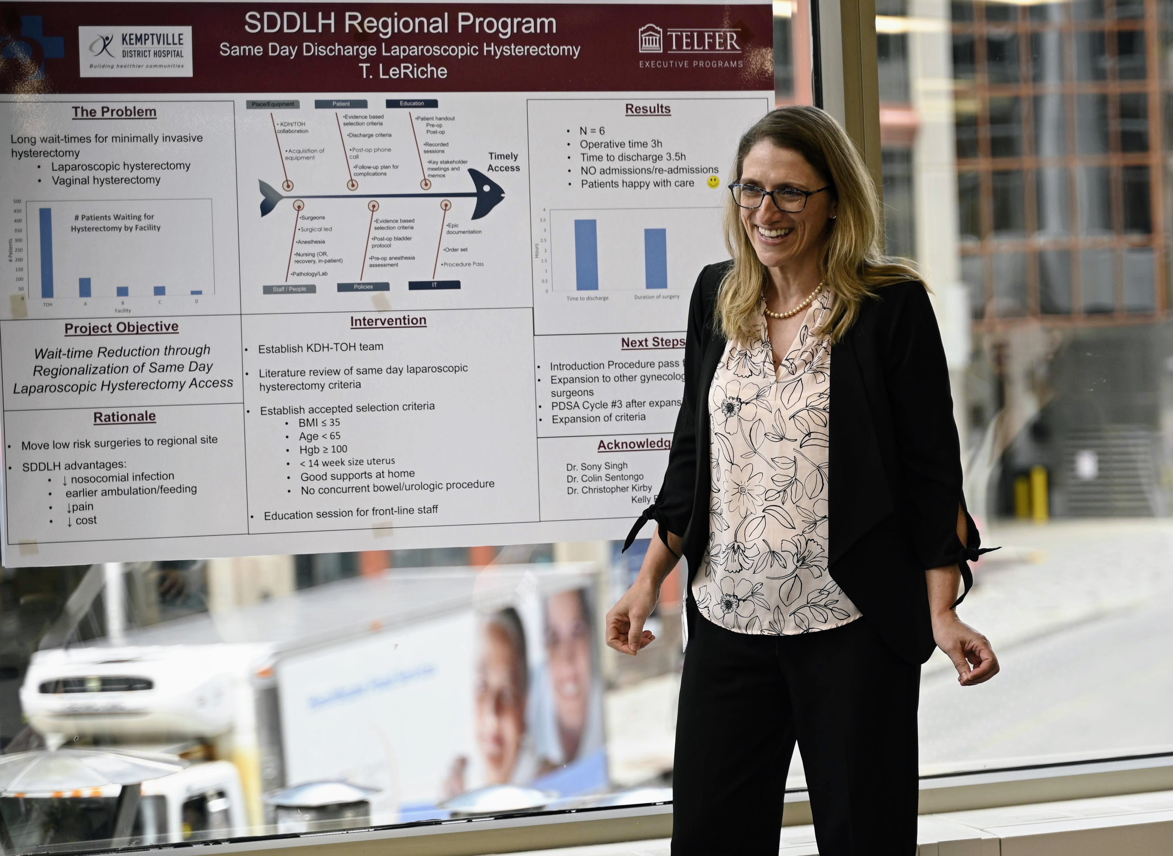 A woman (Dr. Tammy LeRiche) smiles and stands in front of her project poster at the Telfer Centre for Executive Leadership.