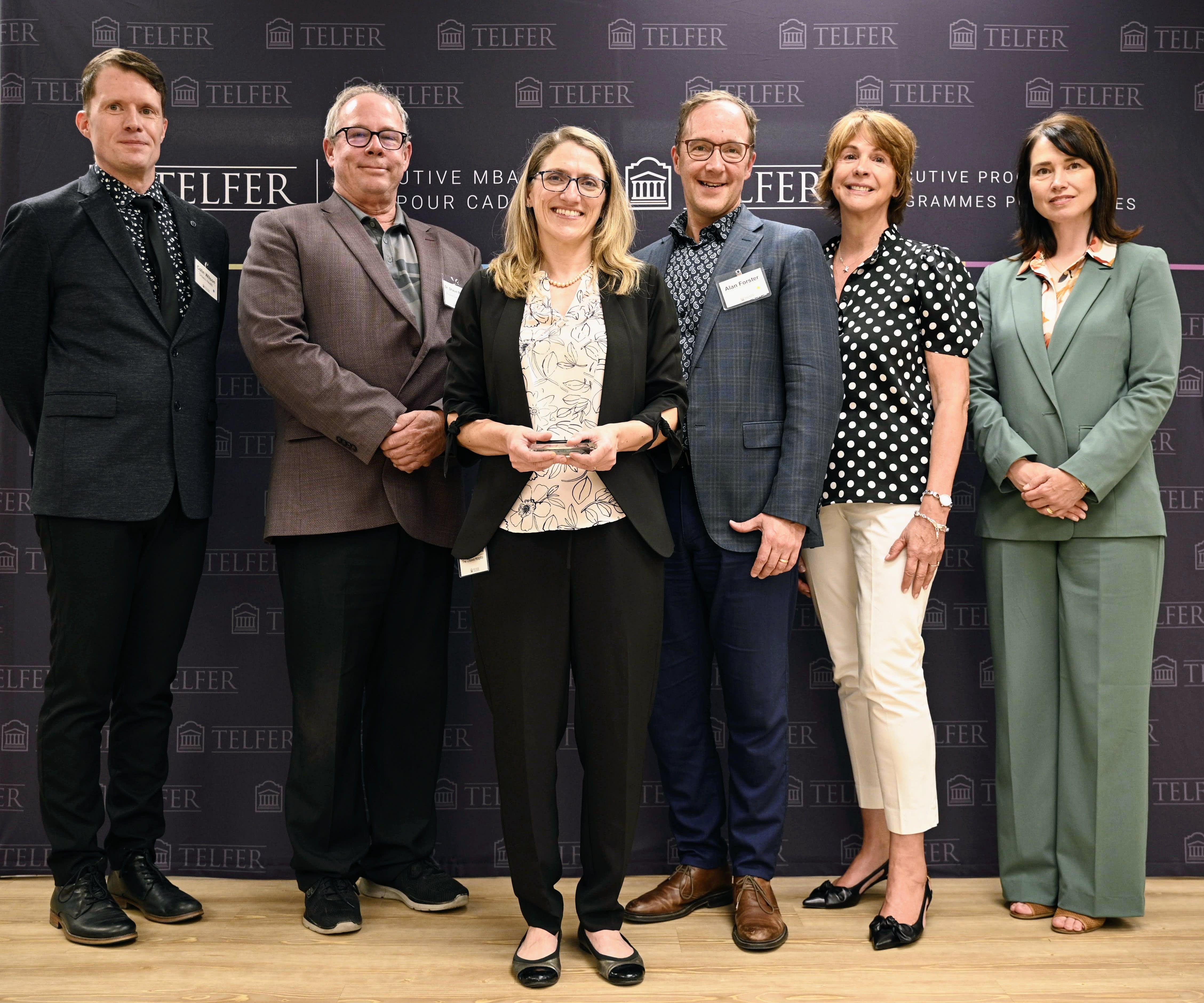 Dr. Colin Wilson, Dr. Shaun McGuire, Dr. Tammy LeRiche, Mary Yates, and Samantha Hamilton stand in front of a backdrop at the Telfer Centre for Executive Leadership.