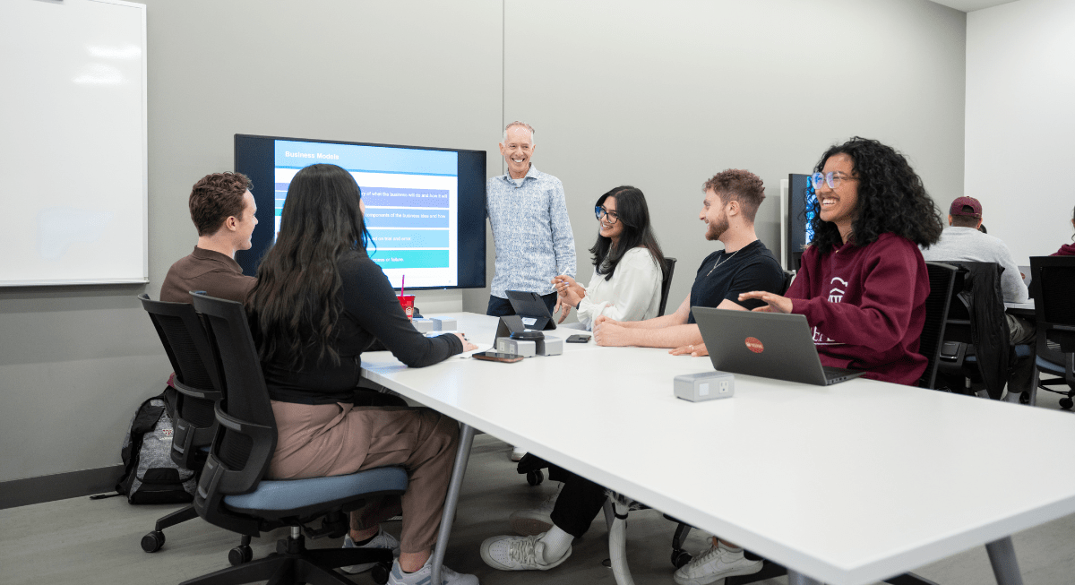 A group of Telfer BCom students and Professor Stephen Daze gathered around a table, smiling and discussing a presentation on a screen.