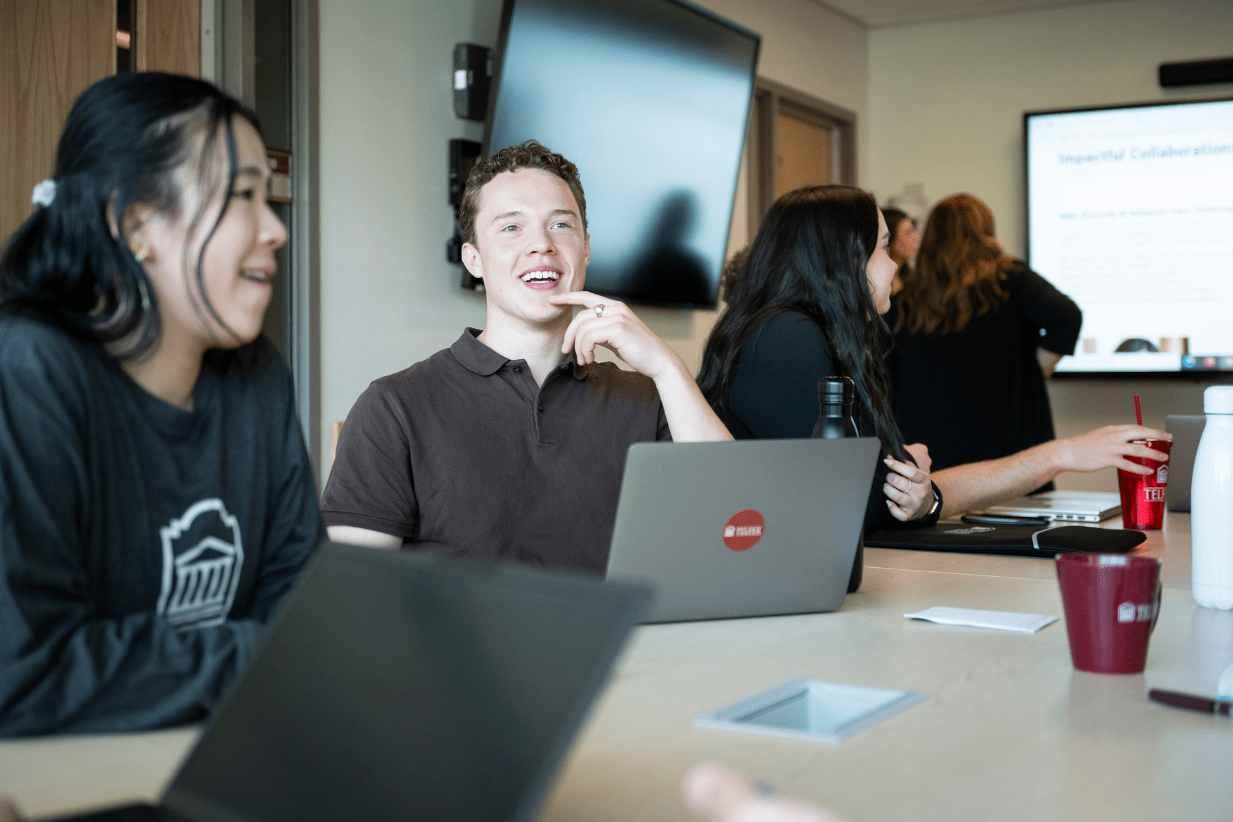 Three Telfer BCom students seated around a table, smiling and engaged in conversation. One student in a brown polo shirt is laughing with a laptop in front of them.