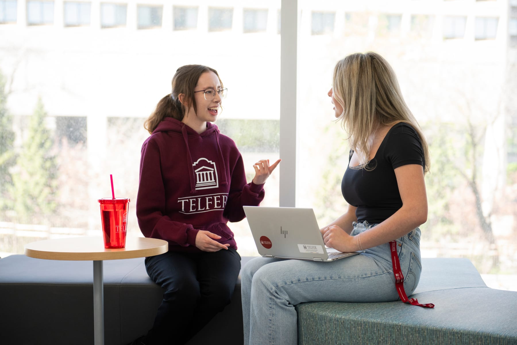 Two female Telfer BCom students facing each other while smiling and engaging in a conversation seated in the student lounge. One student is wearing a garnet Telfer hoodie. The other student has a laptop open on her lap.