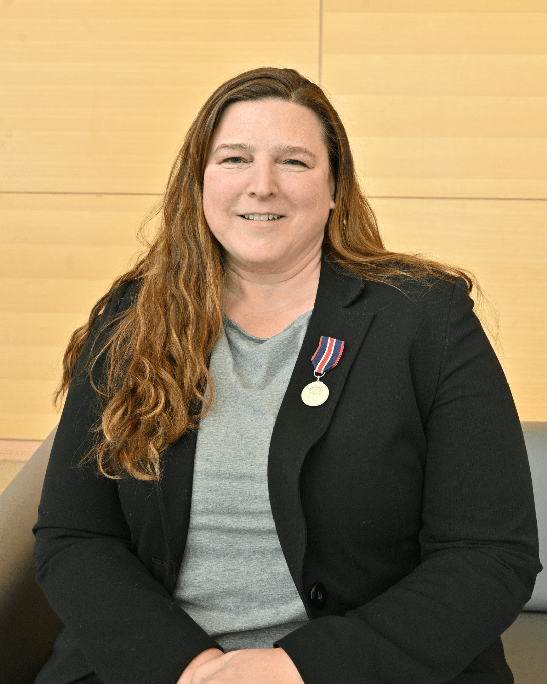 A woman in a suit jacket with a medal pinned to her blazer sits on a couch.