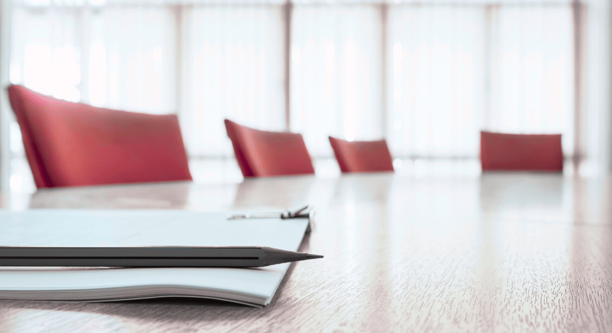 A section of a large table with four empty chairs. A notebook and pencil sits on the table.