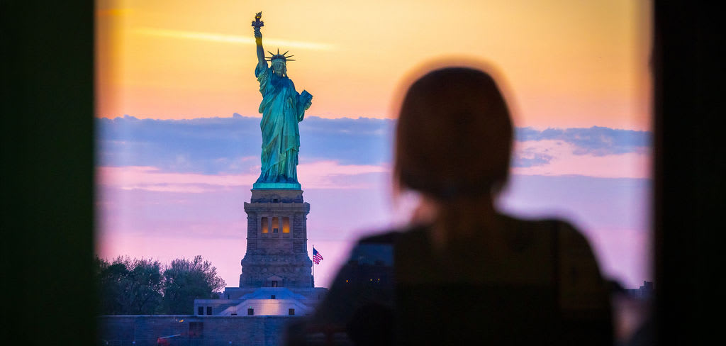 person looking out window at the Statue of Liberty in the distance
