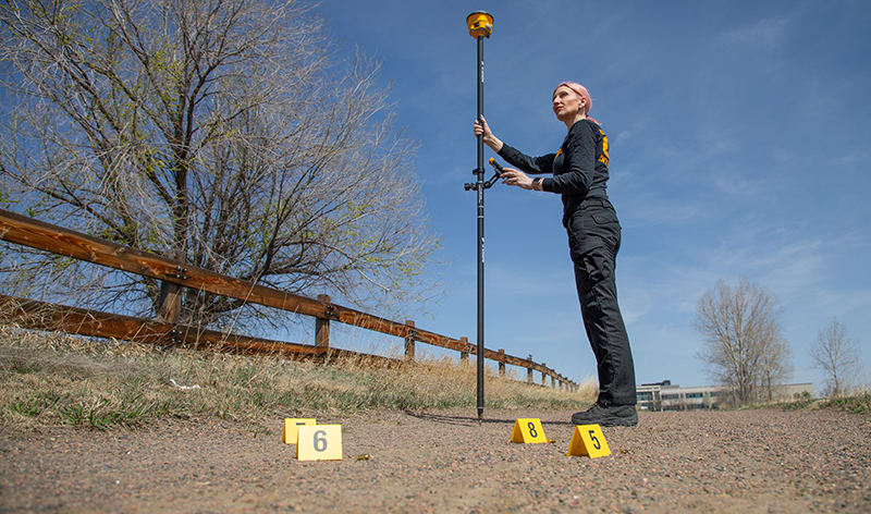 A female crime investigator with a GNSS receiver standing near crime scene evidence markers on the ground.