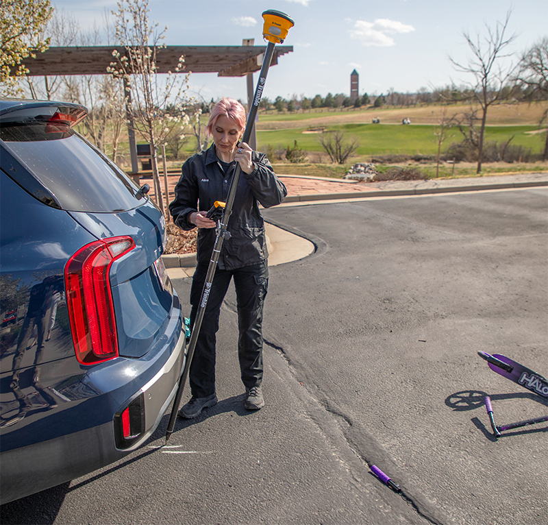 A female crash scene investigator uses a GNSS receiver tilt to document a scooter collision; a blue car is in the background.