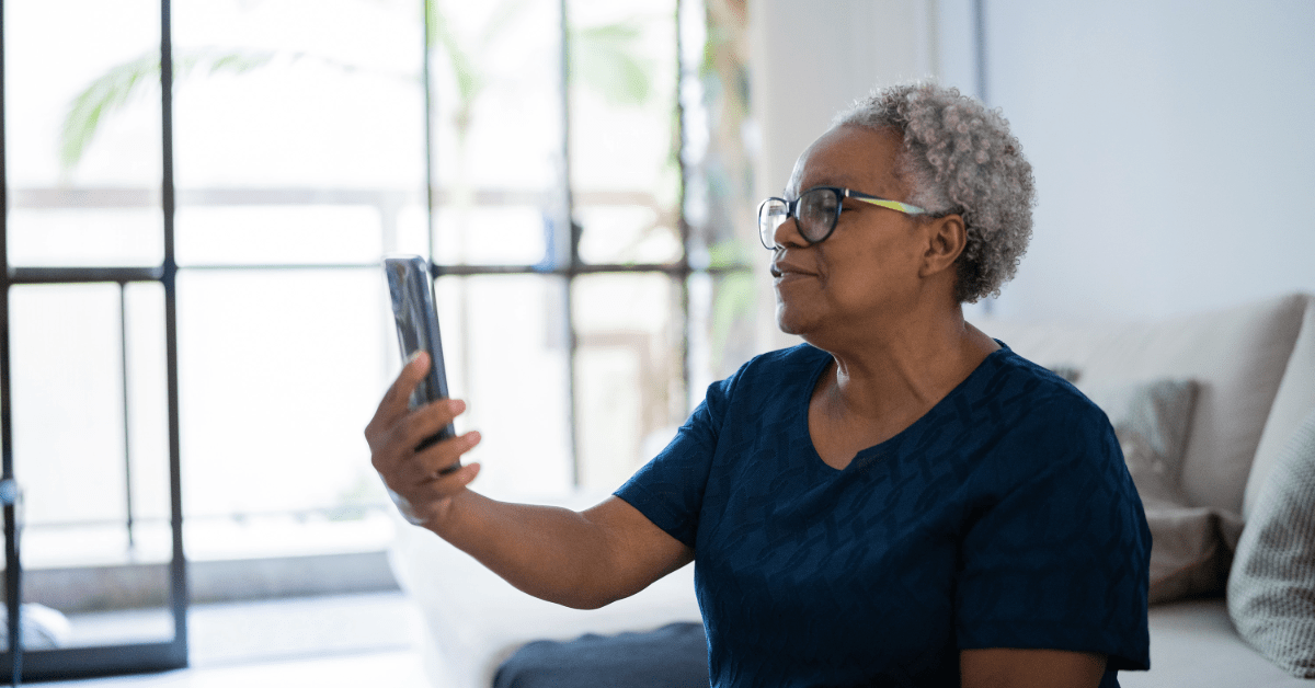 An older woman sitting on a couch, holding a phone up to her face for a telehealth visit