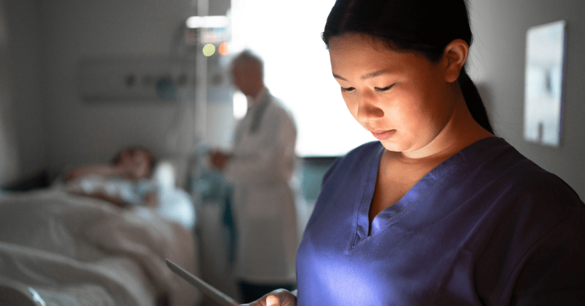 A female nurse in a hospital, looking down at an iPad while in a patient's room