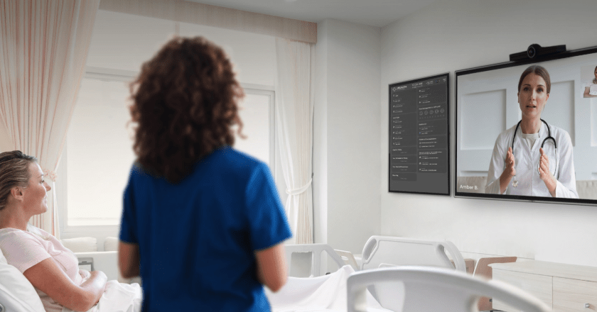 A nurse and a patient looking at a TV screen with another nurse on it and a device with notes on it to the left