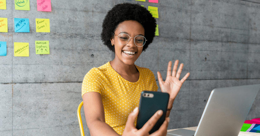 A woman at her desk, holding her phone and waving at a video call with a laptop in front of her, and surrounded by sticky notes