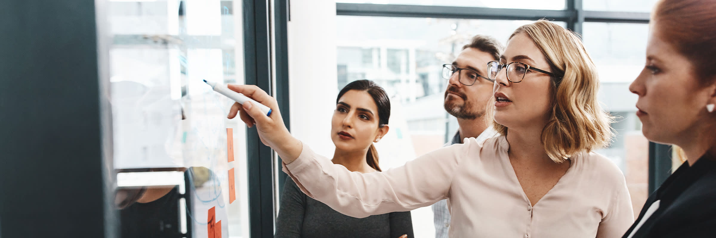 4 people standing in a meeting in front of a whiteboard where one person has a market in her hand and writing on the white board