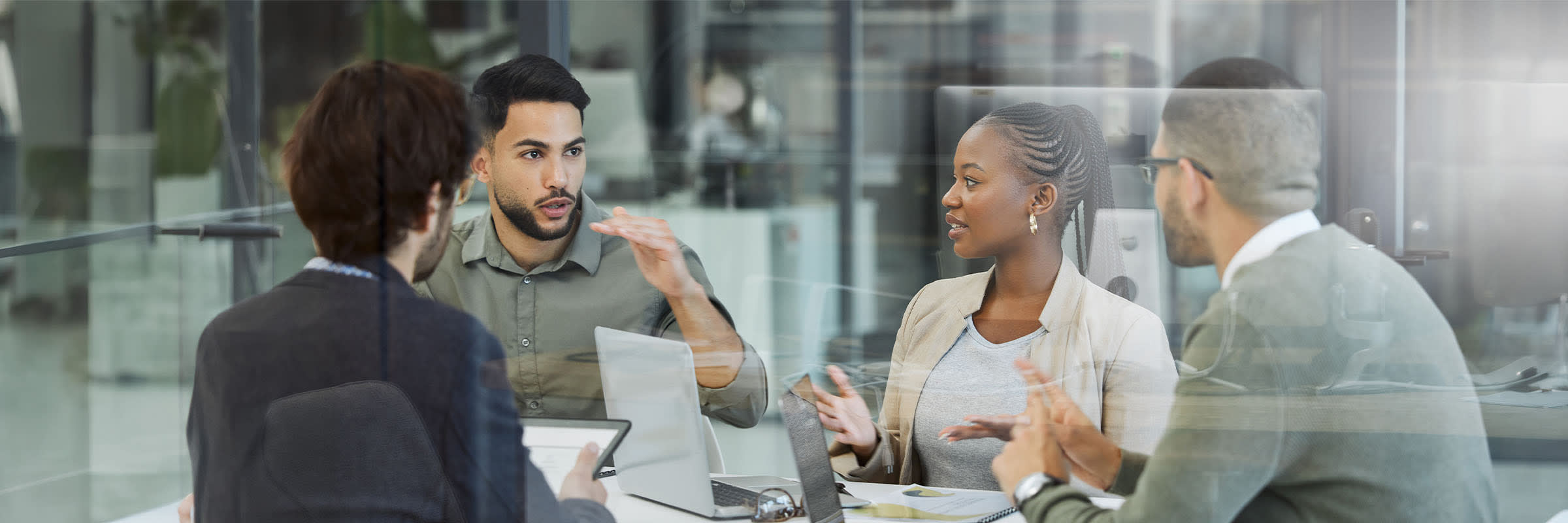 4 people working around a table in a conference room engaging in conversation while on the desk they have their laptops open