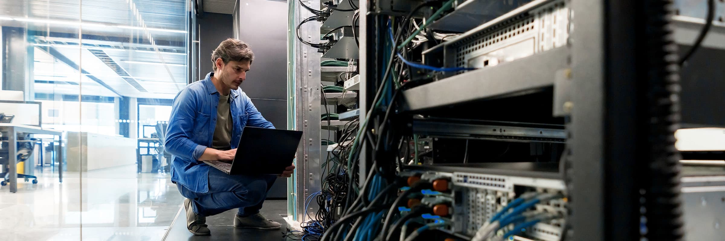 Man crouched balancing a laptop on his right knee while looking at the open rack in a datacentre