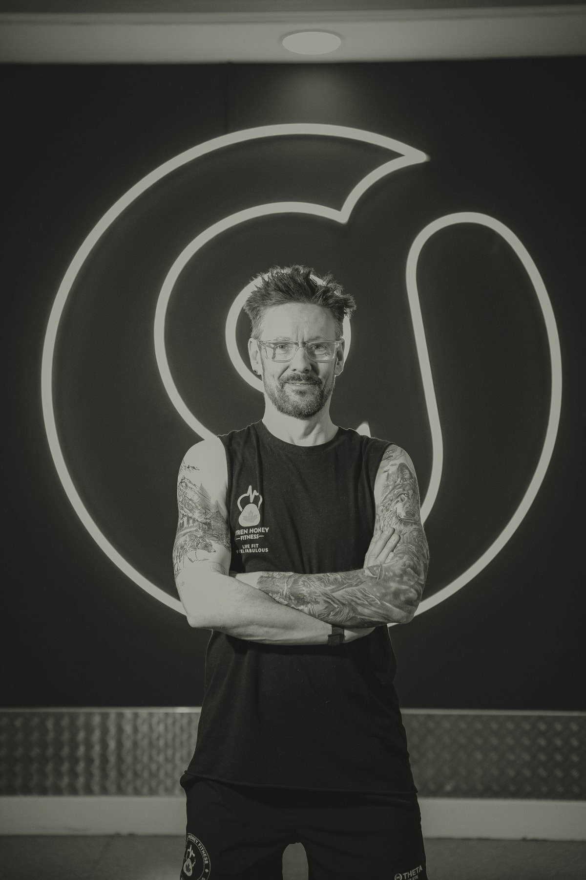 Black and white portrait of Ayren Honey standing with arms crossed in front of the gym's neon logo