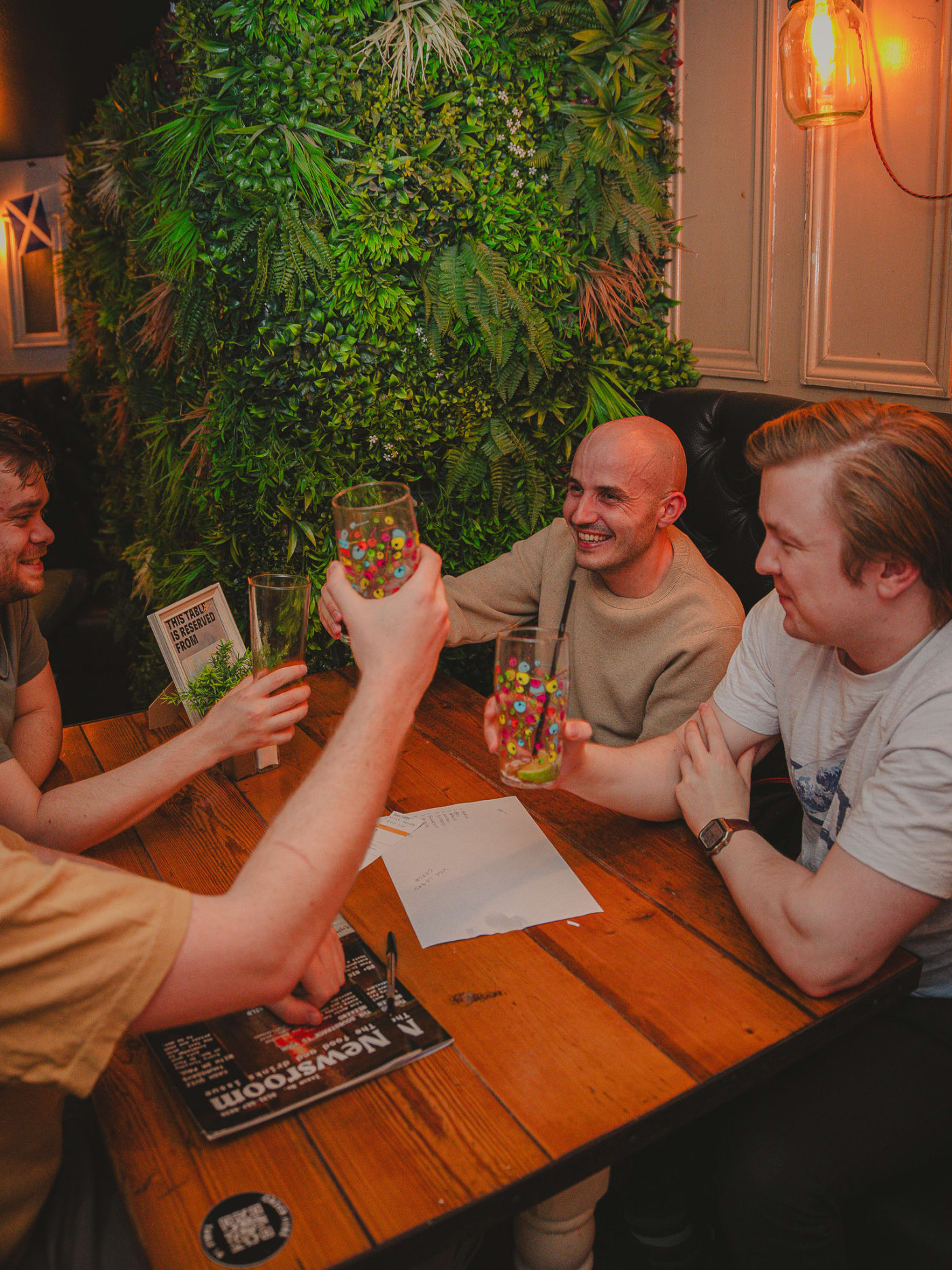 Friends raising their drinks together at a table beside the living plant wall at Newsroom, Edinburgh
