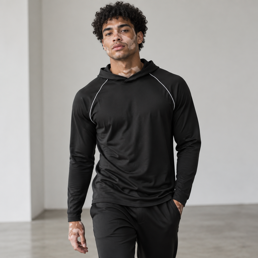 Create a high-fashion editorial photograph featuring a 25-year-old male model with visible vitiligo, showcasing natural and unique irregular patterns primarily on the face, neck, and hands, realistically following the body's anatomy. The model has curly black hair and a fresh, makeup-free appearance. He possesses an athletic build with natural and anatomically correct proportions. The skin texture should be exquisitely detailed, highlighting pores, subtle imperfections, natural tone variations, and a realistic transition between pigmented and depigmented areas due to vitiligo, avoiding any artificial or AI-generated appearances.

The model is dressed in the garment from the reference image, striking a natural walking pose that conveys confidence and elegance. The photograph is captured with a professional camera using a 50mm lens, featuring soft natural lighting, a shallow depth of field, and precise focus on the face and skin texture. The background is a minimalist studio, clean and visually balanced.

The final result should resemble a professional editorial photograph taken by a real fashion photographer, with physically coherent lighting, natural shadows, realistic reflections, correct anatomy, natural proportions, individually defined hair, and an extremely faithful representation of vitiligo. Avoid any plastic skin appearance, excessive smoothing, CGI effects, illustration-like qualities, and artificial features from image generation.