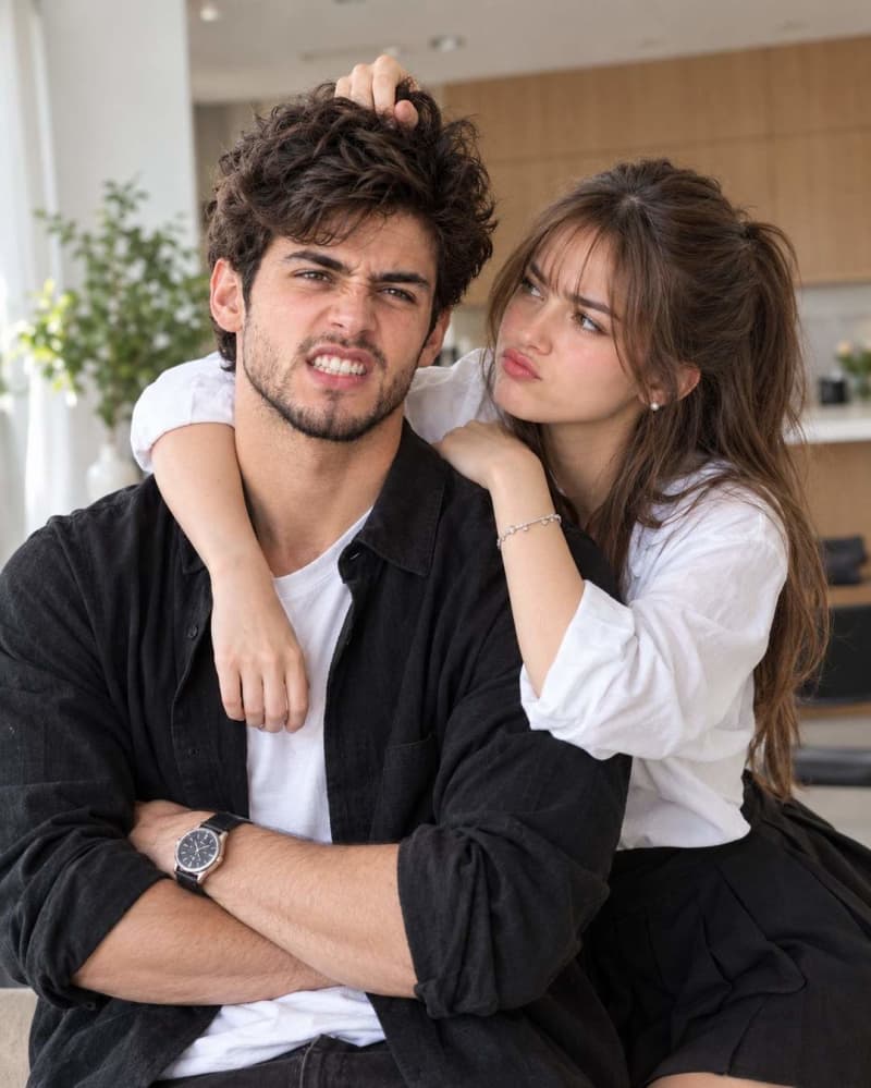 Playful young couple in a bright apartment, the woman pouting while playfully pulling the mans fluffy hair as he scrunches his face in a funny annoyed expression with clenched teeth