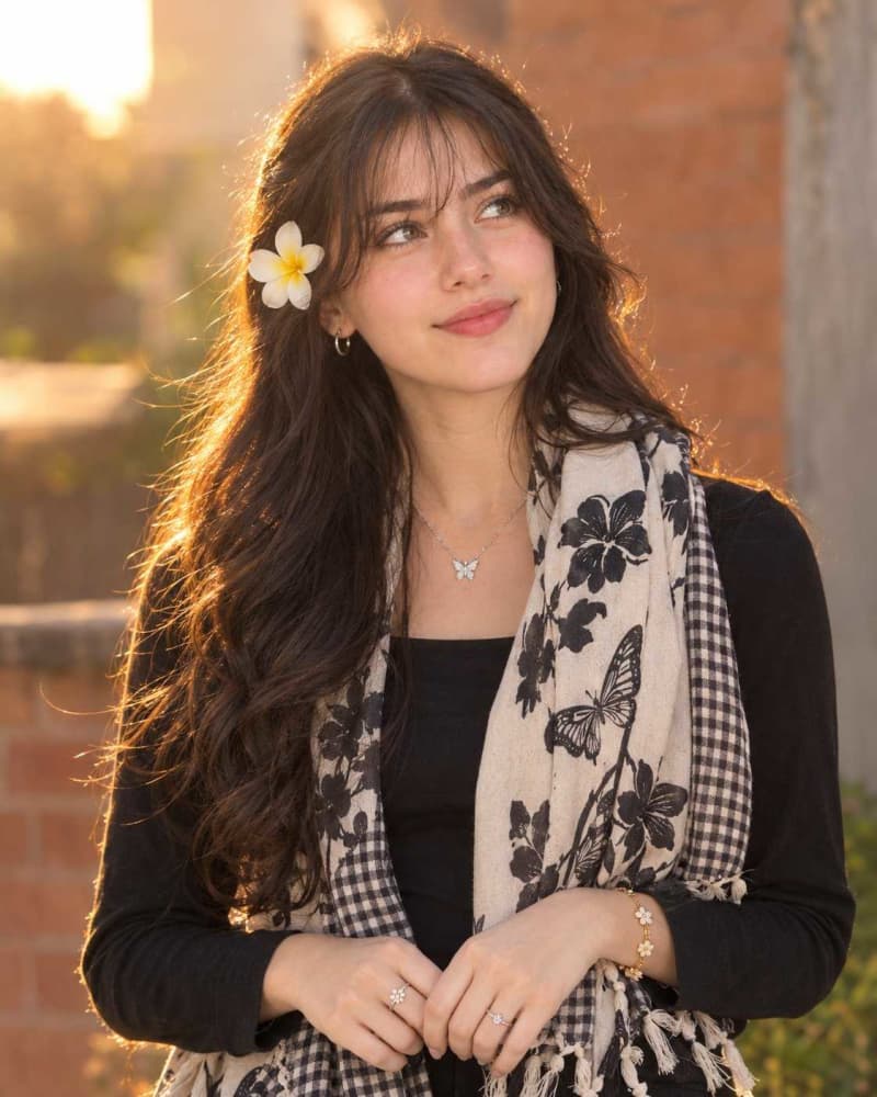 Graceful young woman with long black hair and a white plumeria flower behind her ear, wearing a black top and a cream butterfly print scarf in warm golden hour light