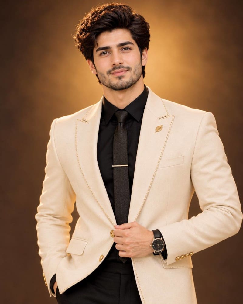 Handsome man with voluminous black hair wearing a cream ivory blazer with gold stitching over a black shirt and tie, standing against a warm golden studio backdrop